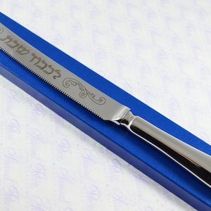 May include: A silver bread knife with a blue box. The knife has a serrated edge and is engraved with the Hebrew words "L'chaim" and a decorative design.