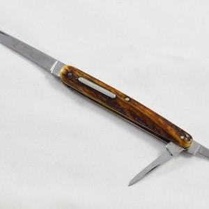 May include: A vintage pocket knife with a polished steel blade and a brown and tan handle. The knife has two blades extended, one long and one short. The handle has a decorative metal inlay.