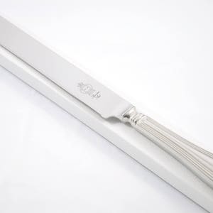 Può includere: Un coltello da torta d'argento con un manico scanalato e una lama appuntita. Il coltello è su una superficie bianca.