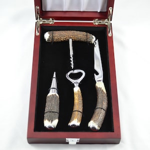 May include: A wine accessory set housed in a dark wood box with a black velvet interior. The set includes a corkscrew, a bottle stopper, a bottle opener, and a foil cutter, all featuring textured brown antler handles with polished metal accents.