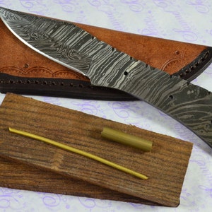 May include: A Damascus steel knife with a brown leather sheath and a wooden handle. The knife is on a white background with a purple pattern.