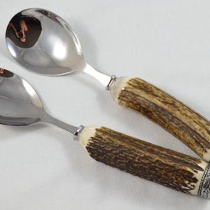 Op de afbeelding: Een set van twee saladeservies van roestvrij staal met geweihandvatten. De handvatten zijn lichtbruin van kleur met een donkerbruin patroon. De servies hebben een gepolijste afwerking.