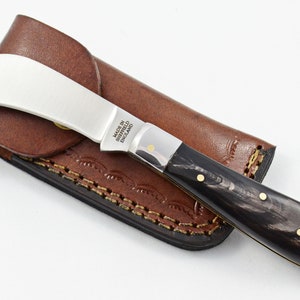 Puede incluir: Un pequeño cuchillo plegable plateado con un mango de cuerno negro y una funda de cuero. El cuchillo tiene una hoja curva y el mango tiene detalles dorados. La hoja está grabada con "Made in Sheffield England".