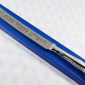 May include: A silver bread knife with an ornate handle and a serrated blade. The blade is engraved with the Hebrew words "ברוך אתה" which translates to "Blessed are you". The knife is in a blue box.