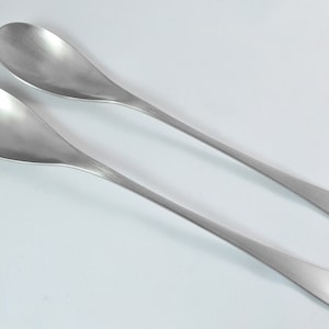 May include: A set of two silver salad serving utensils. The utensils are long and slender with a rounded spoon on one end and a fork with two tines on the other end.
