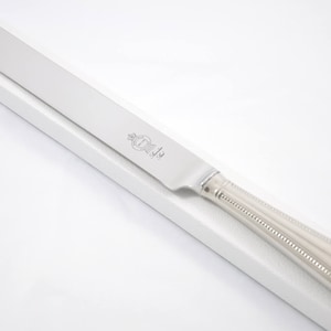 Può includere: Un coltello da torta d'argento con un manico perlato e una lama appuntita. Il coltello è appoggiato su una superficie bianca.