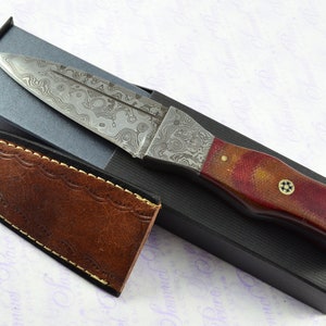 May include: A Damascus steel dagger with a red and brown wood handle. The dagger is in a black box with a brown leather sheath.