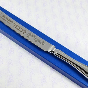 May include: A silver bread knife with a serrated edge and a decorative handle. The knife is engraved with the Hebrew words "L'Chaim" which means "To Life!"