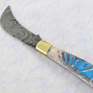 May include: A curved blade knife with a patterned steel blade and a blue, white, and brown marbled handle. The handle has a gold colored metal accent.