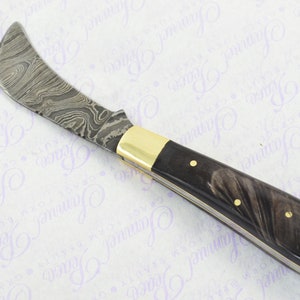 May include: A folding pocket knife with a curved blade made of patterned steel. The handle is black with a marbled pattern and gold accents.