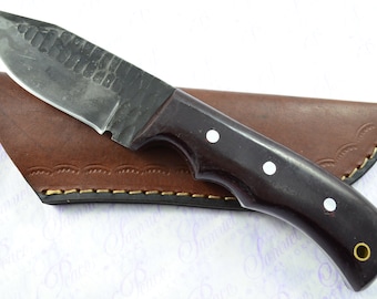 May include: A handmade knife with a dark wood handle and a leather sheath. The blade is made of steel and has a hammered finish.