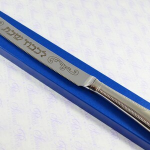May include: A silver bread knife with a blue box. The knife has a serrated edge and a beaded handle. The blade is engraved with Hebrew text and a floral design.