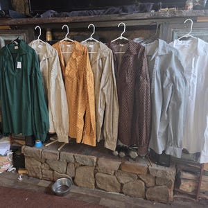 Pode incluir: Uma coleção de sete camisas de manga comprida penduradas em cabides de plástico branco. As camisas apresentam várias cores e padrões, incluindo verde sólido, riscas, amarelo com uma estampa geométrica, castanho com padrão, azul claro e branco.