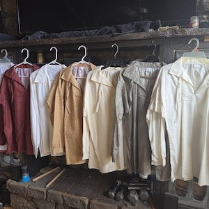 May include: A collection of eight long-sleeved shirts hanging on white and black plastic hangers. The shirts feature a variety of solid colors including red, white, mustard yellow, and grey, with some displaying subtle patterns and textures.