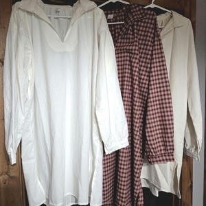 May include: Three long-sleeved shirts hanging on white plastic hangers. The first is a solid white shirt with a V-neck collar. The second is a maroon and white plaid shirt with a collar. The third is a solid off-white shirt with a collar.
