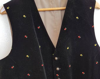 May include: Black velvet vest with a V-neck and small embroidered floral details in red and yellow. The vest has a button-up front with silver-toned buttons and two front pockets. The vest is hanging on a wooden hanger.