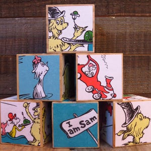 May include: A set of colorful wooden blocks featuring illustrations from the children's book "The Cat in the Hat". The blocks depict characters like the Cat in the Hat, Thing 1, Thing 2, and the Fish.