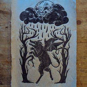 May include: A black ink block print on textured off-white paper featuring a winged, horned creature standing between two bare trees. Above the creature, a dark cloud rains down, and a large, round moon with a face is visible in the upper centre.