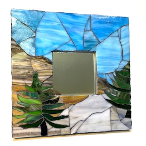 May include: A square mosaic mirror with a blue sky, brown mountains, and a green pine tree. The mirror is framed in a dark brown wood.