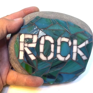 Può includere: Una roccia grigia con un design a mosaico di piastrelle di vetro blu, verde e bianco. La parola "ROCK" è scritta in piastrelle bianche.