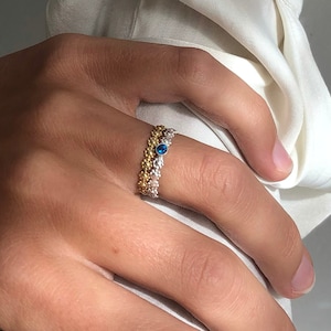 Könnte beinhalten: Ein goldener und silberner Ring mit einem blauen bösen Auge. Der Ring wird am Zeigefinger einer Hand getragen.