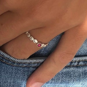 Könnte beinhalten: Ein silberner Ring mit einem kleinen rosafarbenen Stein in der Mitte. Der Ring wird am Mittelfinger einer Hand getragen.