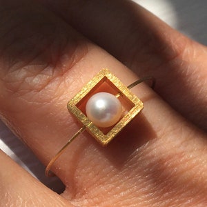 Könnte beinhalten: Ein goldener Ring mit einem quadratischen Rahmen, der eine weiße Perle in der Mitte hält.