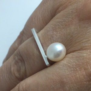 Könnte beinhalten: Ein silberner Ring mit einer einzelnen weißen Perle. Der Ring hat ein schlichtes, modernes Design mit einem geraden Band und einer Perle, die dezent versetzt ist.