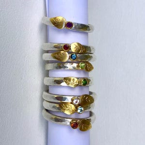 May include: A stack of silver rings with gold leaf accents and colorful gemstones. Each ring features a hammered silver band, a gold leaf design, and a small, round gemstone in various colors, including purple, red, blue, and green.
