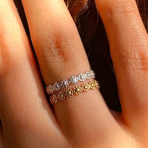 Könnte beinhalten: Zwei zarte Ringe an einem Finger. Der obere Ring ist silber mit einem floralen Design. Der untere Ring ist gold mit einem ähnlichen floralen Design.
