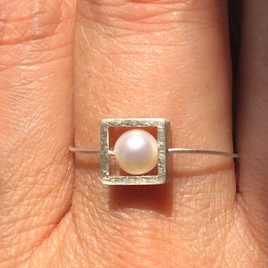Könnte beinhalten: Ein silberner Ring mit einem quadratischen Rahmen, in dem eine weiße Perle sitzt.