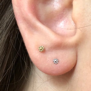 Könnte beinhalten: Nahaufnahme eines Ohres mit zwei kleinen Ohrringen. Der obere Ohrring ist golden mit einem Blumenmuster. Der untere Ohrring ist silber mit einem runden Stein.