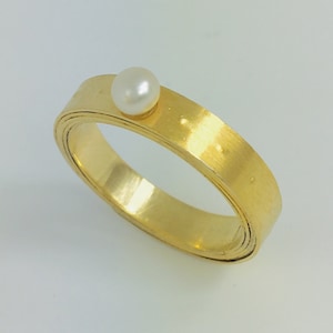 Puede incluir: Un anillo de oro con una sola perla blanca en la parte superior. El anillo está hecho de múltiples bandas de oro, creando un efecto de capas.