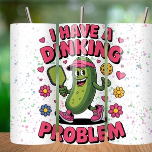 May include: A white cylindrical tumbler with a metal straw featuring a cartoon green pickle wearing a pink visor and sneakers, holding a pickleball paddle. The text reads I HAVE A DINKING PROBLEM in pink letters, surrounded by colorful flowers and hearts.