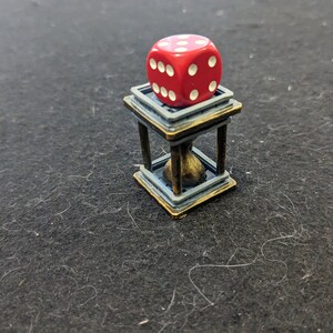May include: A red die with white dots sits atop a miniature hourglass-shaped stand. The stand has a blue and gold color scheme, with a small, round object inside. The die shows the number six.