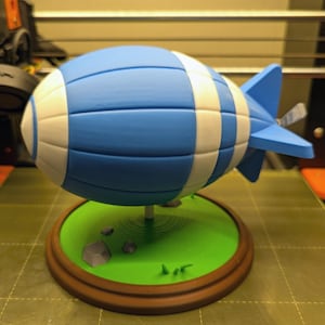May include: A blue and white blimp with a propeller on the back, sitting on a green base with brown trim.