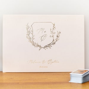 May include: A white guest book with a gold foil design featuring a shield with the initials "M + O" surrounded by a wreath of leaves. The text "Melanie & Oysten 18.06.2022" is printed below the design.
