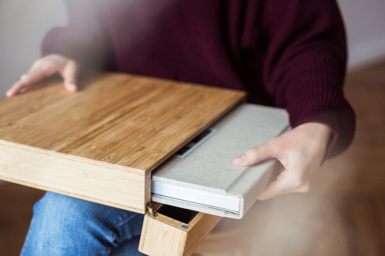 Pode incluir: Uma caixa de madeira clara com um livro a ser retirado. A caixa &eacute; feita de bambu e tem uma tampa articulada. O livro tem uma capa cinza claro e p&aacute;ginas brancas. A pessoa est&aacute; vestindo um su&eacute;ter bord&ocirc; e jeans azul.