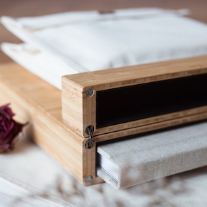 Pode incluir: Uma caixa de madeira com interior preto, uma bolsa de tecido branco e um livro com capa de linho. A caixa &eacute; feita de madeira clara e tem detalhes met&aacute;licos. Uma rosa seca est&aacute; em primeiro plano.