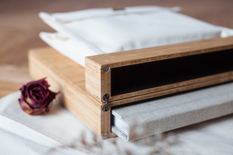 Pode incluir: Uma caixa de madeira com interior preto, uma bolsa de tecido branco e um livro com capa de linho. A caixa &eacute; feita de madeira clara e tem detalhes met&aacute;licos. Uma rosa seca est&aacute; em primeiro plano.