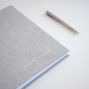 May include: A light gray linen-covered photo album with the names "BIANCA & DANIEL" in gold lettering. A silver pen rests beside the album on a white surface, along with a few decorative stickers.