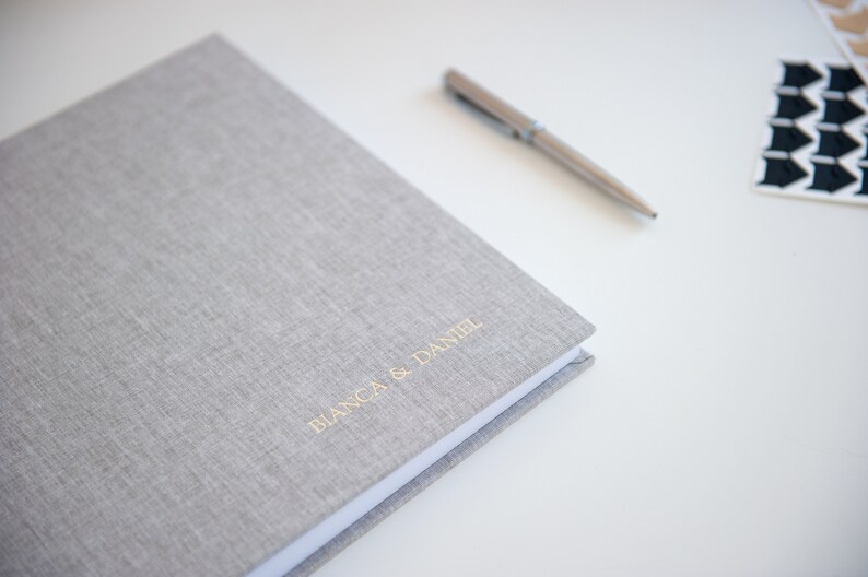 May include: A light gray linen-covered photo album with the names "BIANCA & DANIEL" in gold lettering. A silver pen rests beside the album on a white surface, along with a few decorative stickers.