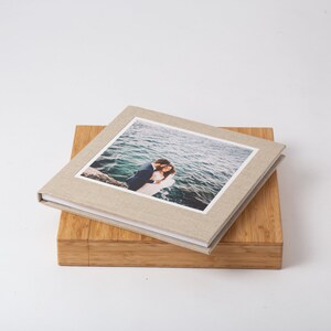 Pode incluir: Um &aacute;lbum de fotos quadrado de linho bege com uma fotografia de um casal abra&ccedil;ado &agrave; beira-mar. O &aacute;lbum repousa sobre uma caixa de madeira, criando um efeito em camadas. O fundo &eacute; branco liso.