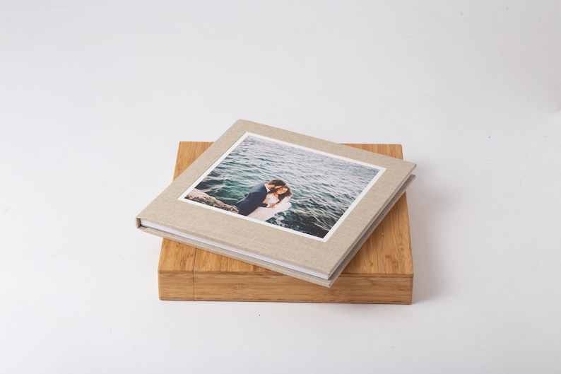 Pode incluir: Um &aacute;lbum de fotos quadrado de linho bege com uma fotografia de um casal abra&ccedil;ado &agrave; beira-mar. O &aacute;lbum repousa sobre uma caixa de madeira, criando um efeito em camadas. O fundo &eacute; branco liso.