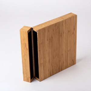 Pode incluir: Uma caixa retangular de bambu parcialmente aberta, com tampa articulada. A caixa &eacute; castanho claro com gr&atilde;o vertical, revelando um interior preto. Em fundo branco.