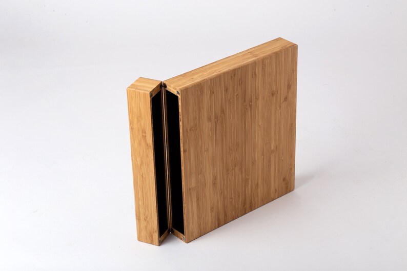 Pode incluir: Uma caixa retangular de bambu parcialmente aberta, com tampa articulada. A caixa &eacute; castanho claro com gr&atilde;o vertical, revelando um interior preto. Em fundo branco.