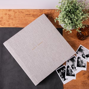 May include: A light gray linen-covered photo album with the names "Olivia + Jeremiah" in gold lettering. The album is next to several black and white photographs on a wooden surface. A small potted plant and a glass candle holder are also present.