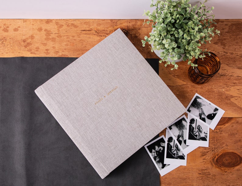 May include: A light gray linen-covered photo album with the names "Olivia + Jeremiah" in gold lettering. The album is next to several black and white photographs on a wooden surface. A small potted plant and a glass candle holder are also present.