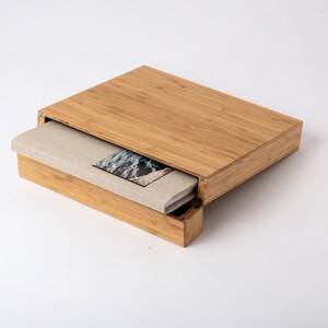 Pode incluir: Uma caixa de bambu natural com tampa deslizante, contendo um livro com capa de linho. O livro apresenta uma fotografia de paisagem na capa. A caixa e o livro est&atilde;o sobre um fundo branco.
