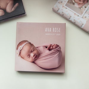 Può includere: Un album fotografico rosa con il testo "AVA ROSE" e "MARCH 21st 2020" sulla copertina. L'album presenta una foto di una bambina che dorme in un fasciatoio rosa.